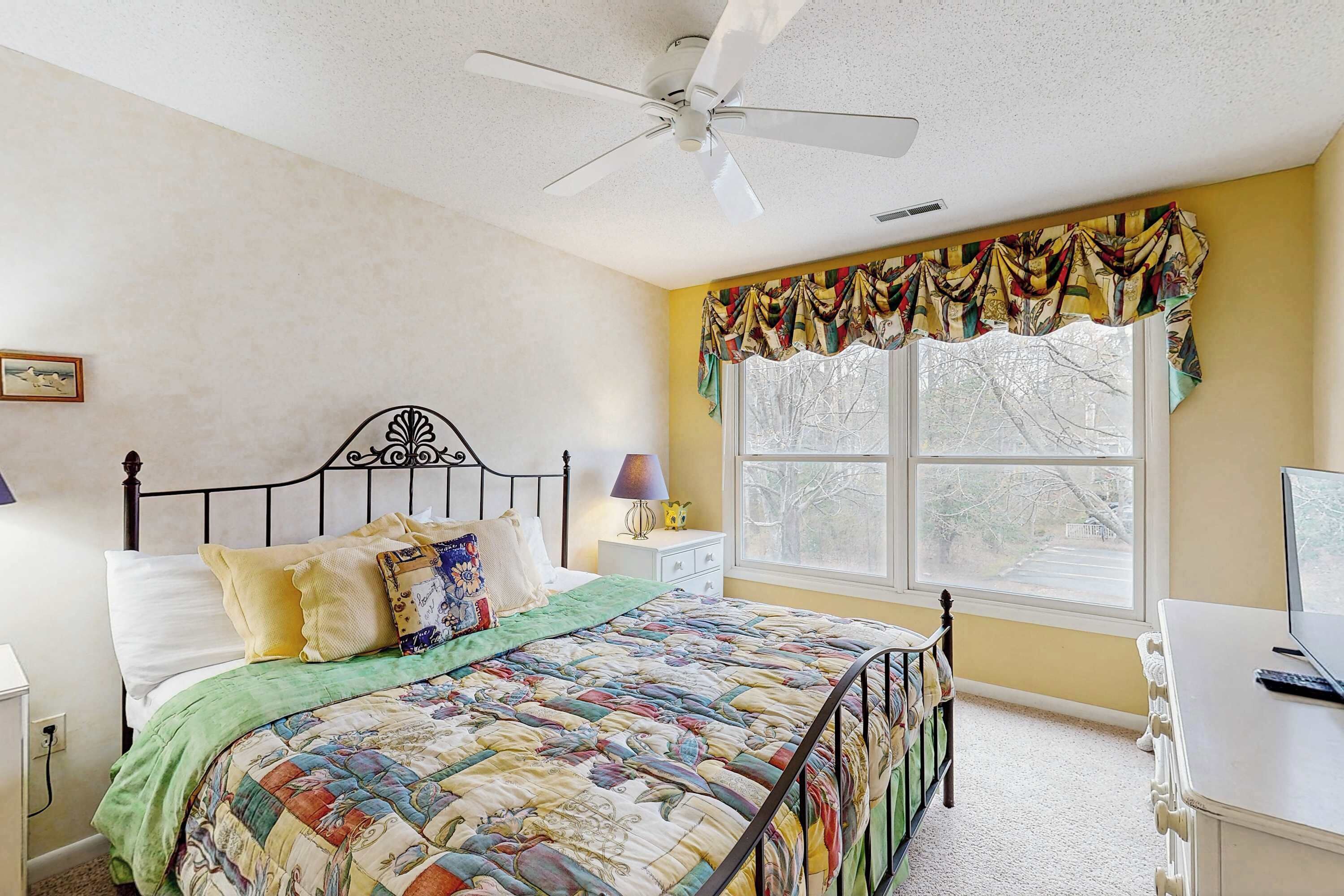 Ein Schlafzimmer mit einem Bett, einem Deckenventilator, einem Fenster mit Vorhängen, einem Nachttisch mit Lampe und einem Schreibtisch mit Computer.