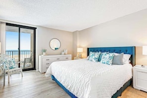 Une chambre d’hôtel avec un grand lit, un balcon avec vue, un miroir rond et une commode blanche.