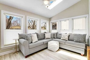 A room with a sectional sofa, a round ottoman, and large windows with views of trees and houses. - Bayberry Woods condo W/ shared pool, high-speed WiFi & central AC - near beach (Bethany Beach)