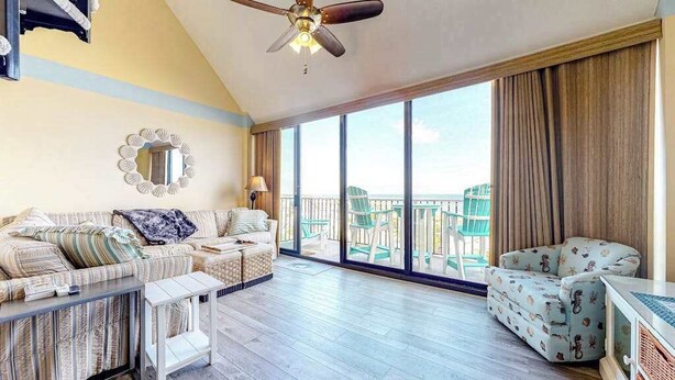 Ein geräumiger Balkon mit Deckenventilator, einer Couch, einem gemusterten Sessel und einem großen Fenster mit Meerblick.