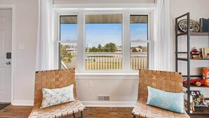 A room with two wicker chairs, a window with a view of a fence and houses, and a bookshelf with various items. - Dog-friendly town of Fenwick home with guest suite, WiFi, & elevator (Fenwick Island)