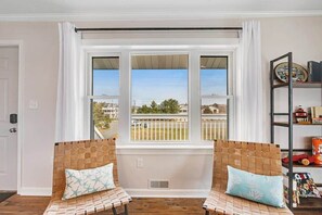 A room with two wicker chairs, a window with a view of a fence and houses, and a bookshelf with various items. - Dog-friendly town of Fenwick home with guest suite, WiFi, & elevator (Fenwick Island)