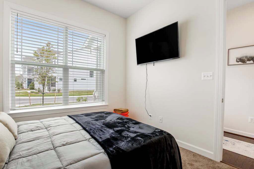 Une chambre à coucher avec un lit, un téléviseur fixé au mur, une fenêtre avec des stores et une porte.