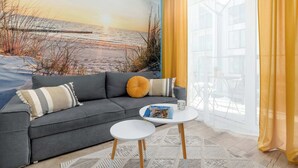 A living room with a grey sofa, a small white table, a beach mural on the wall, and yellow curtains. - Cozy Apartment | Air Conditioning | Balcony | Parking. (Ustronie Morskie)