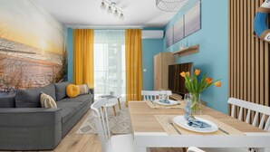 A modern living room with a grey sofa, white chairs, a wooden dining table, and a large painting of a sunset. - Cozy Apartment | Air Conditioning | Balcony | Parking. (Ustronie Morskie)