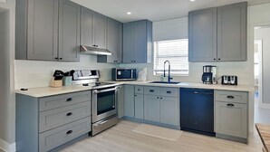 A modern kitchen with grey cabinets, stainless steel appliances, and a window with blinds. - Casa Playa Serena | Remodeled, Volleyball Net + Pet-Friendly (St. Augustine)