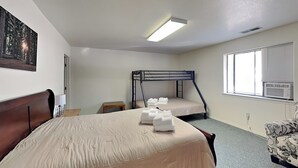 Ein Hotelzimmer mit einem großen Bett, einem kleineren Bett, einem Schreibtisch, einem Stuhl, einem Bild an der Wand und einem Fenster mit Jalousien.