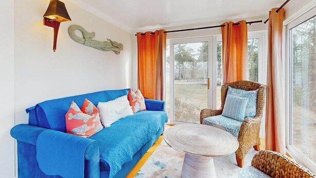 Habitació amb un sofà blau, cadires de vimet, una taula rodona, una làmpada penjada a la paret i una porta de vidre que dona a l'exterior.