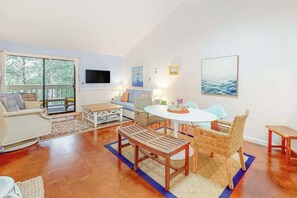 A spacious living area with a large window, a sofa, a dining table, and wicker furniture.