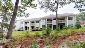 Un edificio de dos pisos con un porche delantero, un pequeño jardín con rocas y arbustos, y un árbol en primer plano.