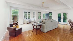 A spacious living room with a large sofa, a coffee table, and a TV. There are multiple windows providing natural light and views of greenery outside. - Dog-friendly historic home with yard, games, grill, children's items, W/D, & AC (Barnstable)