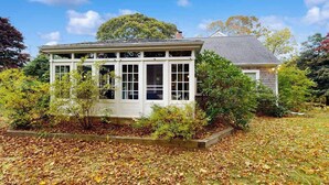 A white house with a covered porch, surrounded by trees and a leaf-covered lawn. - Dog-friendly historic home with yard, games, grill, children's items, W/D, & AC (Barnstable)