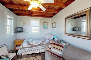 A bedroom with a wooden ceiling, a bed with a patterned quilt, a sofa, a bedside table, a lamp, a mirror, and a television.