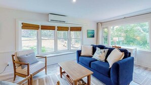 Una sala de estar con un sofá azul, una silla de madera, una mesa de centro y ventanas grandes con persianas.
