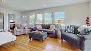 A spacious living room with a grey sofa, a striped armchair, and a wooden coffee table. - Oceanview Buzzards Bay getaway w/ wraparound deck - steps to the Beach (Bourne)
