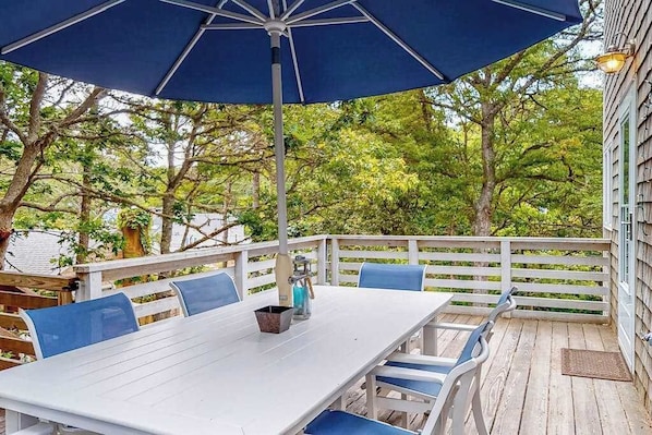 Eine überdachte Terrasse im Freien mit einem weißen Tisch und blauen Stühlen, umgeben von einem Holzdeck und einem weißen Zaun.