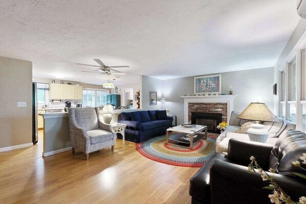 A spacious living room with a fireplace, a dining area, and a kitchen in the background. - Historic Barnstable Village Home w/ Private Yard - Near Beach (Barnstable)