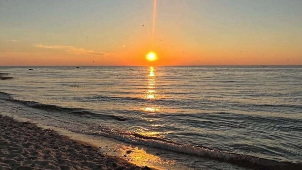 A beach with the sun setting over the water, creating a reflection on the surface.