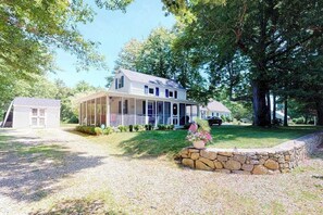 A two-story house with a covered porch, a stone wall, and a small shed in the background. - Welcoming home on Main St. with screened porch & large yard - 1 dog OK (Cotuit)