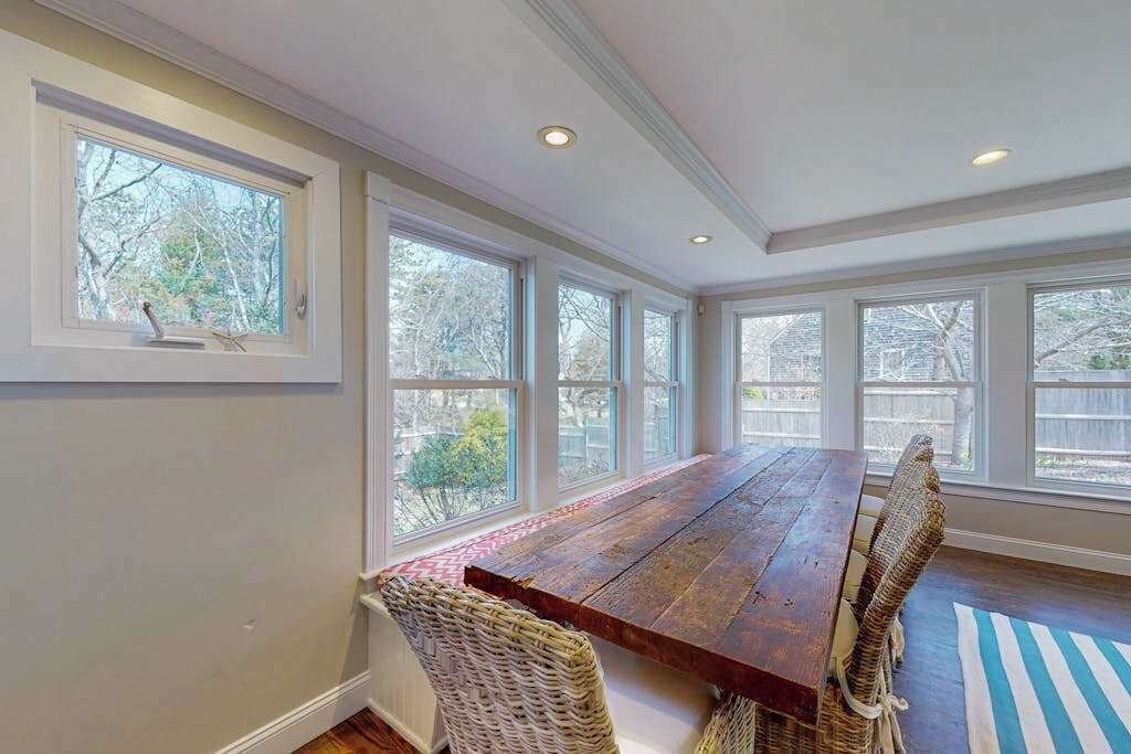 A dining area with a long wooden table, wicker chairs, large windows, and a view of trees.