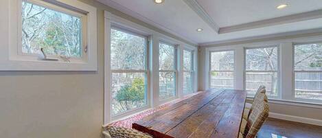 A dining area with a long wooden table, wicker chairs, large windows, and a view of trees.
