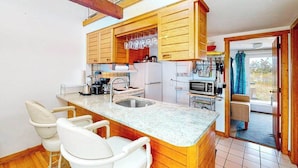 Una cocina con armarios de madera, encimera de granito, un fregadero y un microondas. Hay dos sillas blancas y una mesita.