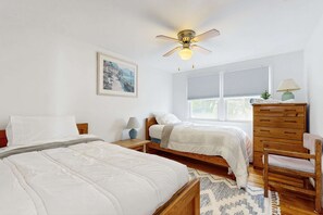 A bedroom with two beds, a ceiling fan, a window with blinds, a dresser, and a lamp.