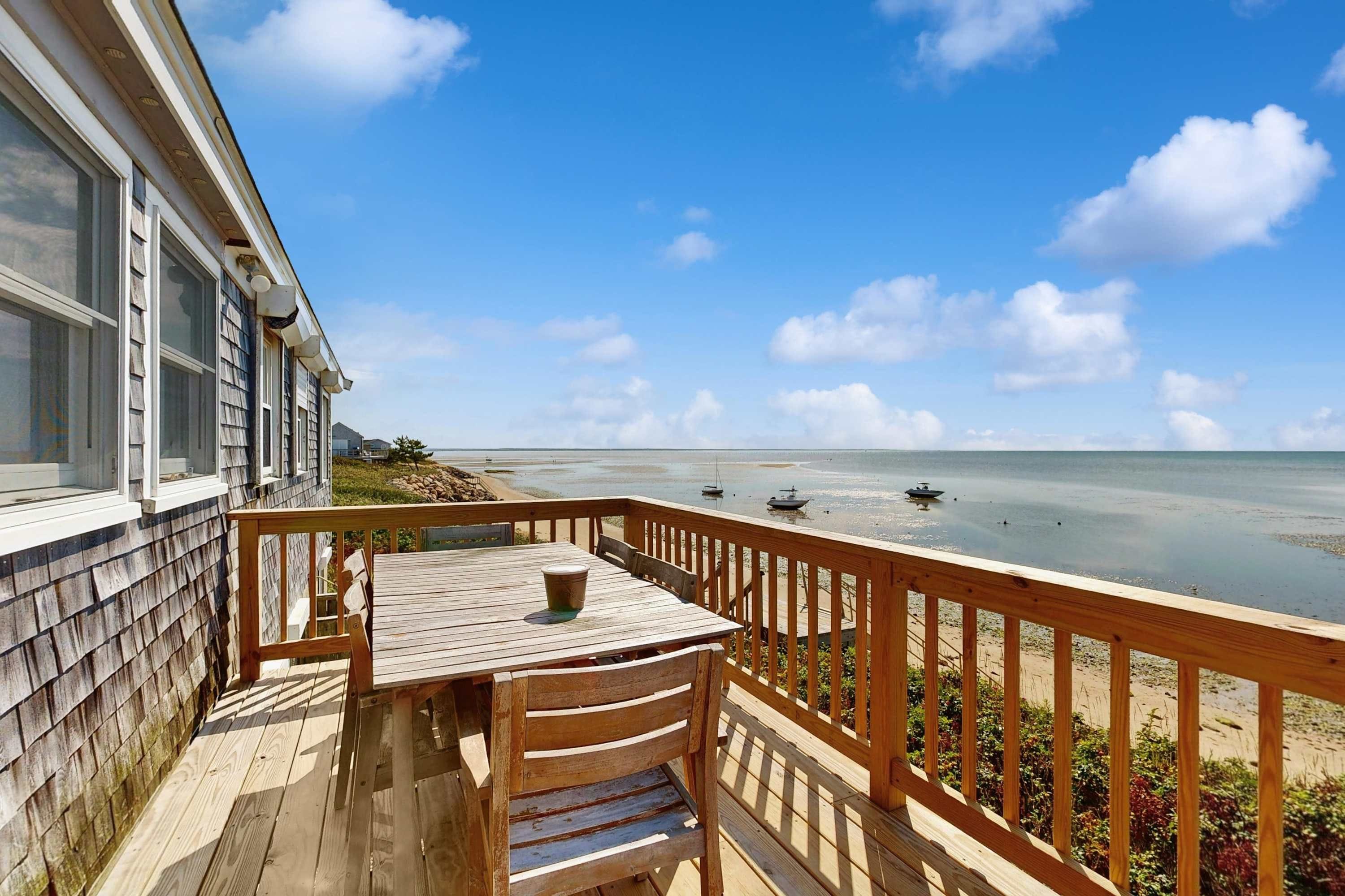A wooden deck with a table and chairs, overlooking a beach with boats and a clear sky.