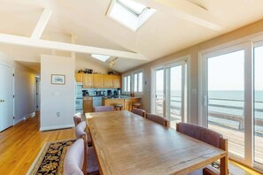 A spacious room with a large wooden dining table, a kitchen area, and a view of the ocean through large windows.