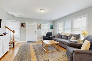 A living room with a sofa, armchair, coffee table, and a staircase. - Newly built bayview home, steps to dock/launch on Great Pond - small dogs OK (Falmouth)