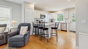 Cocina moderna con armarios blancos, zona de comedor con sofá gris y taburetes de bar, y piso de madera.