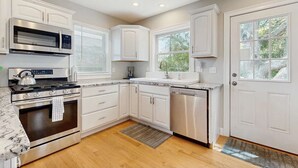 Una cocina con armarios blancos, una estufa de acero inoxidable, lavavajillas y ventana con vista a árboles.