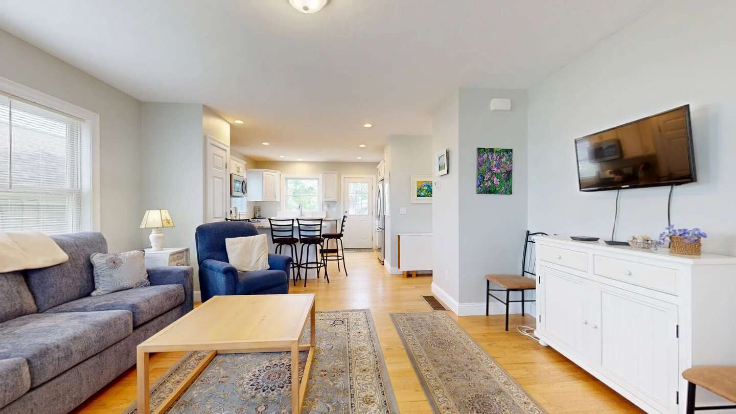 Een moderne woonkamer met een bank, salontafel en televisie. Er is een eethoek met stoelen en een tafel. De keuken is op de achtergrond zichtbaar.