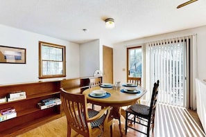 A dining area with a round table, chairs, a bookshelf, and a view through a window.