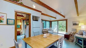 A spacious room with a wooden dining table, chairs, a sofa, and a large window offering a view of greenery.