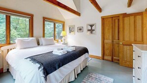 A bedroom with a bed, bedside tables, a lamp, framed pictures, wooden wardrobe, and large windows.
