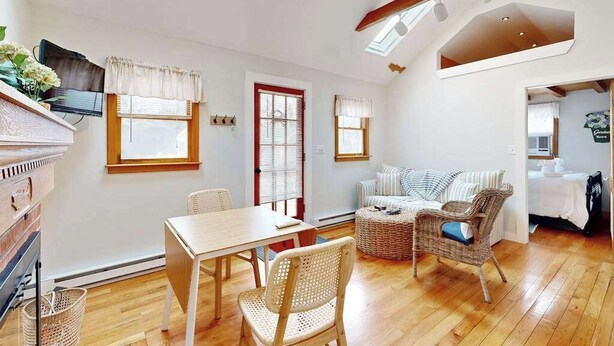 A room with a wooden floor, a dining table with chairs, a sofa, and a television on the wall. - Cottage near Cape Cod National Seashore with firepit, grill, kitchenette, & AC (Eastham)