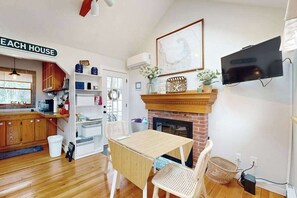 A cozy kitchen with a fireplace, wooden cabinets, a dining table, and a TV mounted on the wall. - Cottage near Cape Cod National Seashore with firepit, grill, kitchenette, & AC (Eastham)