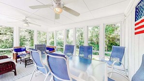 Une véranda avec une grande table, des chaises, des ventilateurs de plafond et une vue sur la verdure.