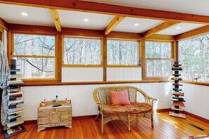 A cozy room with a wooden floor, a wicker chair with a cushion, a small wooden dresser, and a tall bookcase. - Gorgeous woodland home with deck & firepit - walk to ocean bluffs & Rail Trail (Eastham)