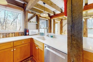 A kitchen with wooden cabinets, a white countertop, a stainless steel dishwasher, and a window with a view of trees. - Gorgeous woodland home with deck & firepit - walk to ocean bluffs & Rail Trail (Eastham)