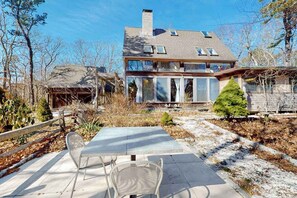 A two-story house with a patio, a small tree, and a wooden fence. - Gorgeous woodland home with deck & firepit - walk to ocean bluffs & Rail Trail (Eastham)