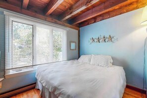 A bedroom with a bed, wooden ceiling beams, a window with blinds, and a wall decoration.