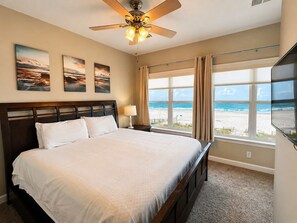 A bedroom with a bed, a ceiling fan, a nightstand, a lamp, and a view of the beach. - Gulf Front | 6 Bedrooms | Spacious West Beach Retreat (Gulf Shores)