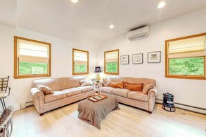 Un salon avec un canapé d’angle beige, une table basse et une lampe. Des tableaux encadrés ornent le mur, et une fenêtre offre une vue sur la verdure.