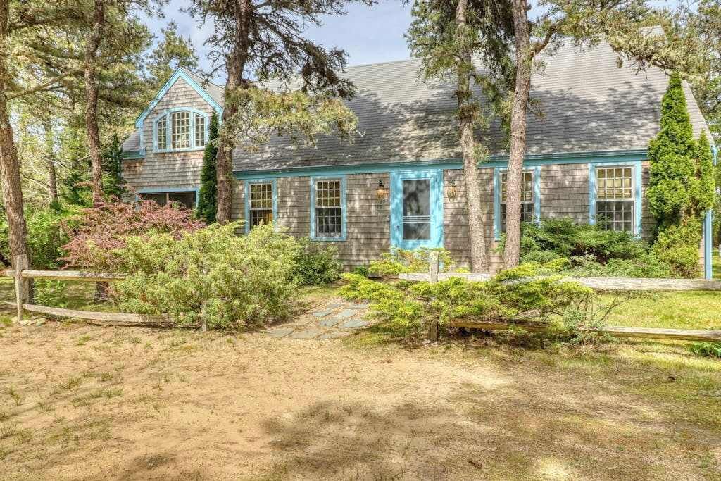 A wooden house with a gabled roof, surrounded by trees and shrubs.