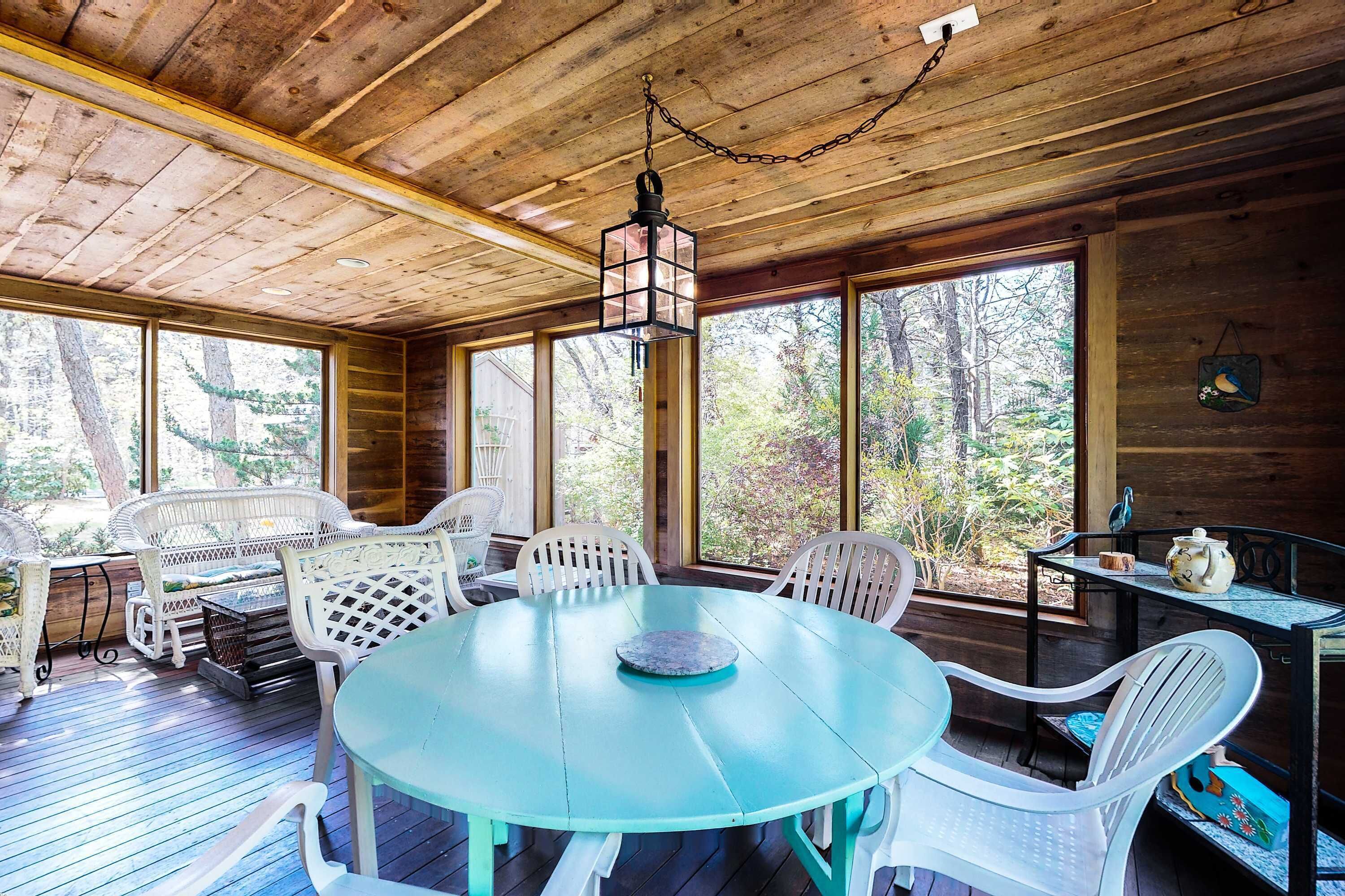 A wooden deck with a round table and white chairs, surrounded by large windows.