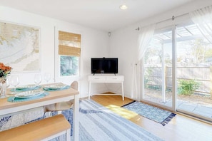 Un coin repas avec une table dressée pour quatre personnes, un téléviseur posé sur un meuble blanc et une grande fenêtre donnant sur l’extérieur.