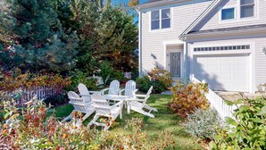 Ein weißes Haus mit Garage, umgeben von einem Garten mit verschiedenen Pflanzen und einer Sitzgelegenheit mit Adirondack-Stühlen.