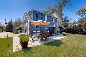 A two-story house with a patio area featuring a table and chairs, a grill, and potted plants.